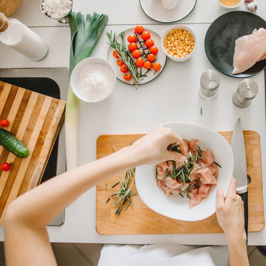 Overhead view of a person preparing raw chicken in a white bowl with rosemary on a wooden cutting board, surrounded by fresh vegetables, spices, and kitchen items on a white countertop.
