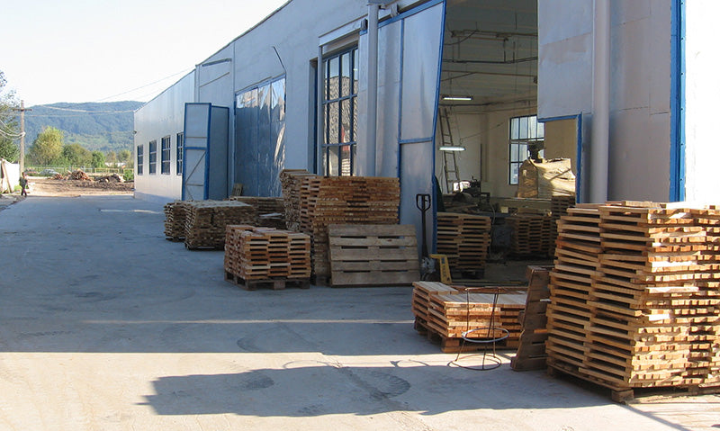 Stacks of wooden pallets arranged outside a large industrial warehouse with open metal doors and a concrete yard.