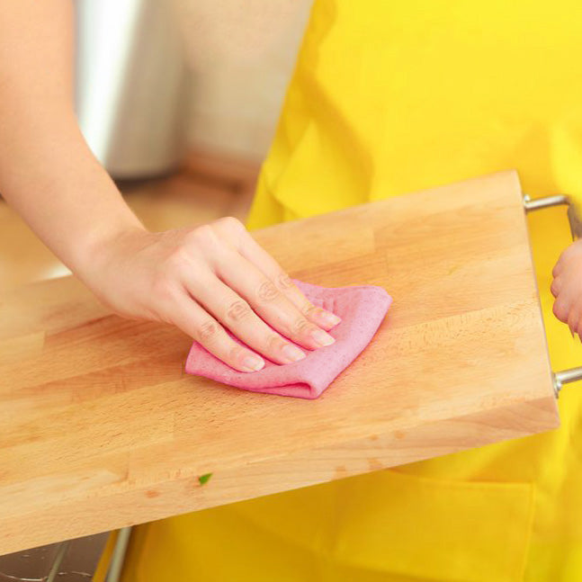 A person wearing a bright yellow apron wipes a beechwood cutting board with a pink cloth in a kitchen setting.