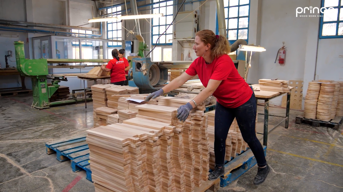 A woman in a red t-shirt and dark pants stands in a woodworking factory, handling stacks of unfinished light-colored wooden blocks on a blue pallet. Another worker is visible in the background near large industrial machines.