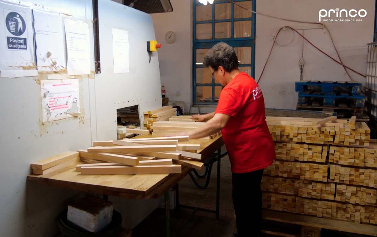 A person in a red t-shirt stands at a workbench in a workshop, handling wooden strips next to a large machine. Stacks of wooden blocks are visible on pallets in the background, with a 'princo' watermark in the top right corner.